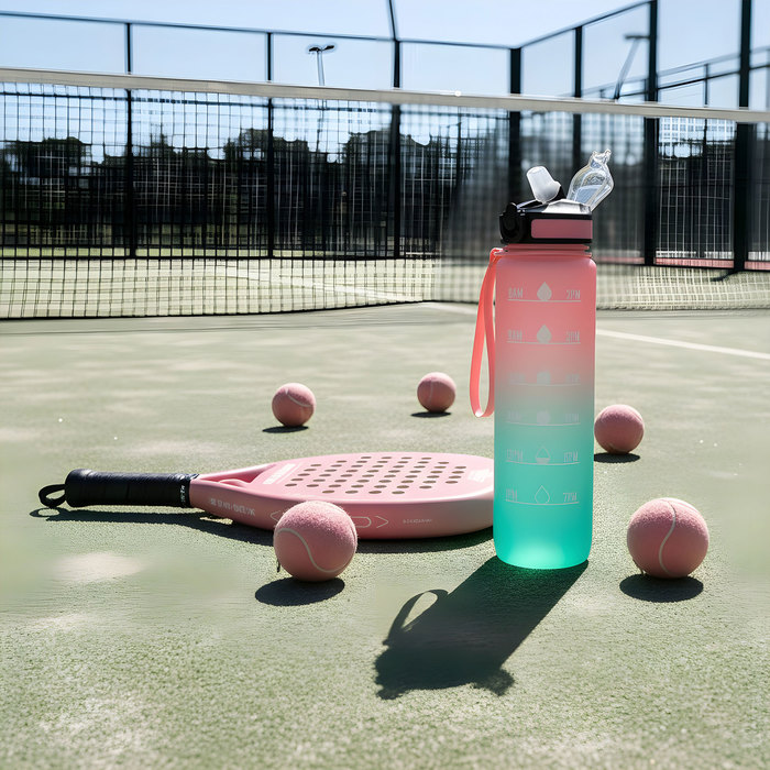 La botella deportiva motivacional GRAF en color rosa/aguamarina con acabado traslucido, con contador de tiempo y medidor y su cinta de transporte en color rosa, apoyada en el piso de una cancha de tenis