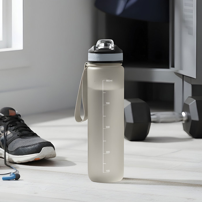 Botella deportiva motivacional NADAL en color gris con acabado frost, con marcador medida, tapa con boquilla rebatible y correa. Apoyada en el piso.