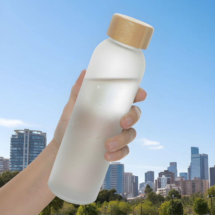Mano sosteniendo Botella sublimable SANDER y al fondo un paisaje citadino, hecha de vidrio resistente con un elegante acabado frost, su tapa de bambú antiderrames, con interior de acero inoxidable.